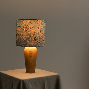 Load image into Gallery viewer, Lamp, lit, sits on natural linen tablecloth, against a soft grey background.  Marbled cotton lampshade in Stone marbling pattern in dark teal and soft lemon, hint of black.  Vase style wooden base turned on Dartmoor
