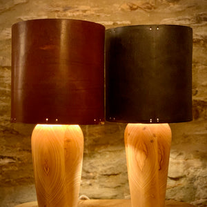 Load image into Gallery viewer, Veg-tanned leather lampshades on curved wooden base, sitting on wooden table with stone wall background.  Leather lampshades in Dark Oak and Dark London Tan colours.  Ash wooden lamp base