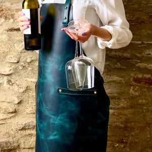 Load image into Gallery viewer, Commissioned ACQUA veg tanned leather apron.  Woman standing against a stone wall backdrop, wearing the apron over a white shirt, holding bottle of wine and two wine glasses.