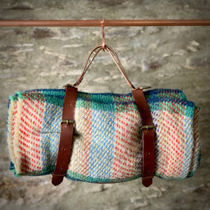 Load image into Gallery viewer, ROCWORX Oak bark veg-tanned leather holder with multi-coloured recycled woollen picnic blanket.  Holder has brass buckles, d-rings and copper rivets, with easy carry leather handle.  Hanging from copper hook on a copper pipe with stone wall in the background.