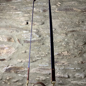 Load image into Gallery viewer, ROCWORX Oak bark veg-tanned leather holder with multi-coloured recycled woollen picnic blanket.  Holder has brass buckles, d-rings and copper rivets, with easy carry leather handle.  Hanging from Navy webbing and leather shoulder strap on copper hook on a copper pipe with stone wall in the background.