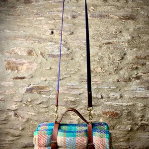 Load image into Gallery viewer, ROCWORX Oak bark veg-tanned leather holder with multi-coloured recycled woollen picnic blanket.  Holder has brass buckles, d-rings and copper rivets, with easy carry leather handle.  Hanging from Navy webbing and leather shoulder strap on copper hook on a copper pipe with stone wall in the background.