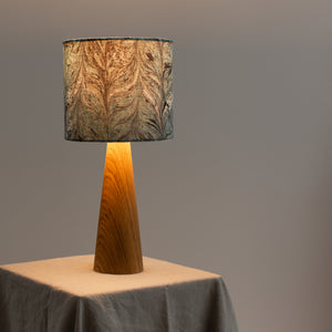 Load image into Gallery viewer, Lamp lit.  Sitting on natural linen tablecloth with soft grey wall background.  Chevron marbling pattern in dark teal and dark green on linen lampshade.  Wooden cone style lamp base.
