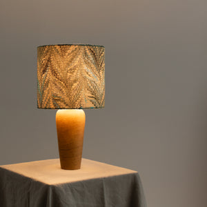 Load image into Gallery viewer, Lamp lit, sitting on a natural linen tablecloth, with soft grey background.  Linen lampshade in Feathers Nonpareil marbling pattern drum shade, in dark green and lemon, with hint of teal.  On a wooden base

