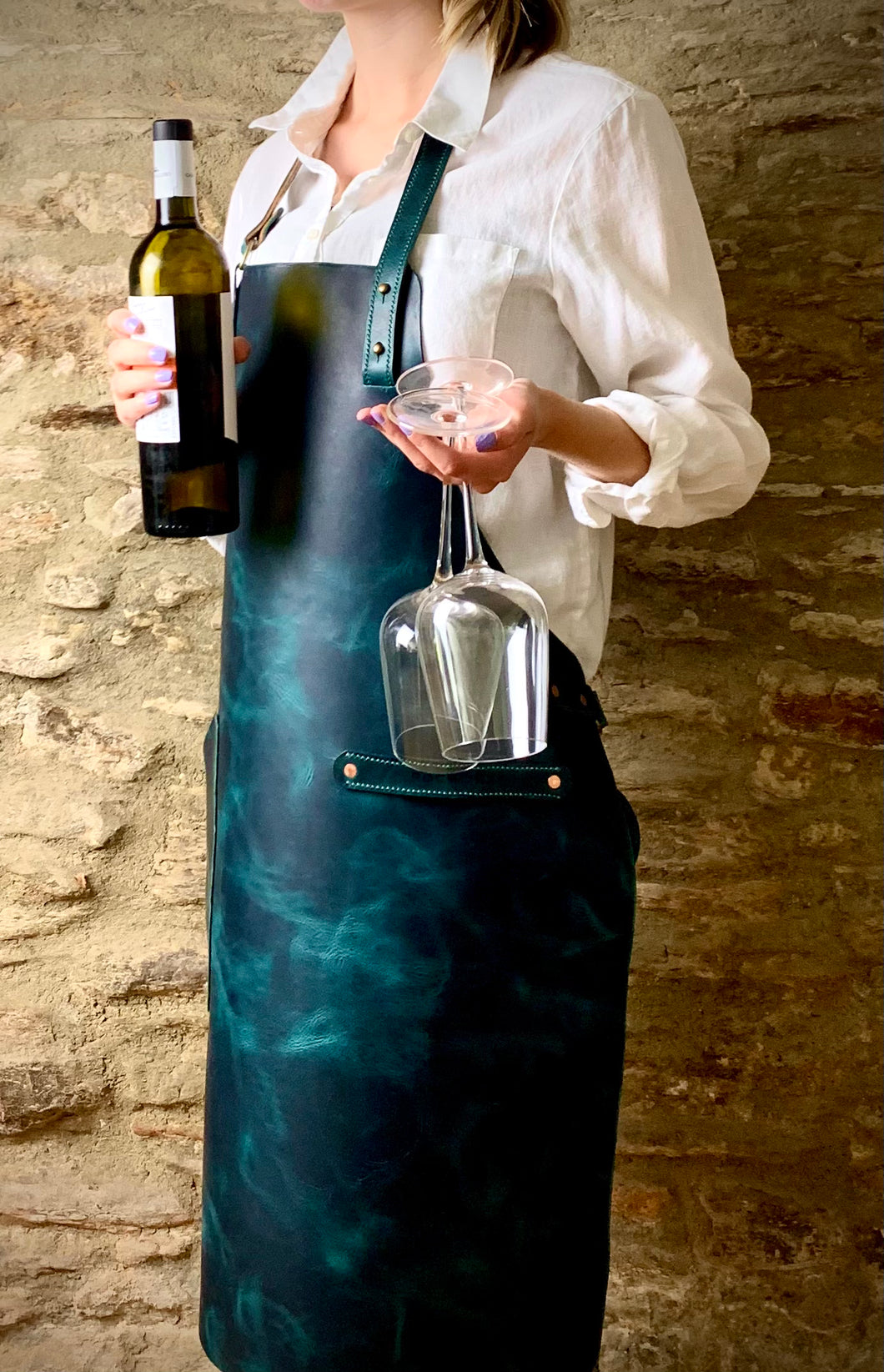 Commissioned ACQUA veg tanned leather apron.  Woman standing against a stone wall backdrop, wearing the apron over a white shirt, holding bottle of wine and two wine glasses.